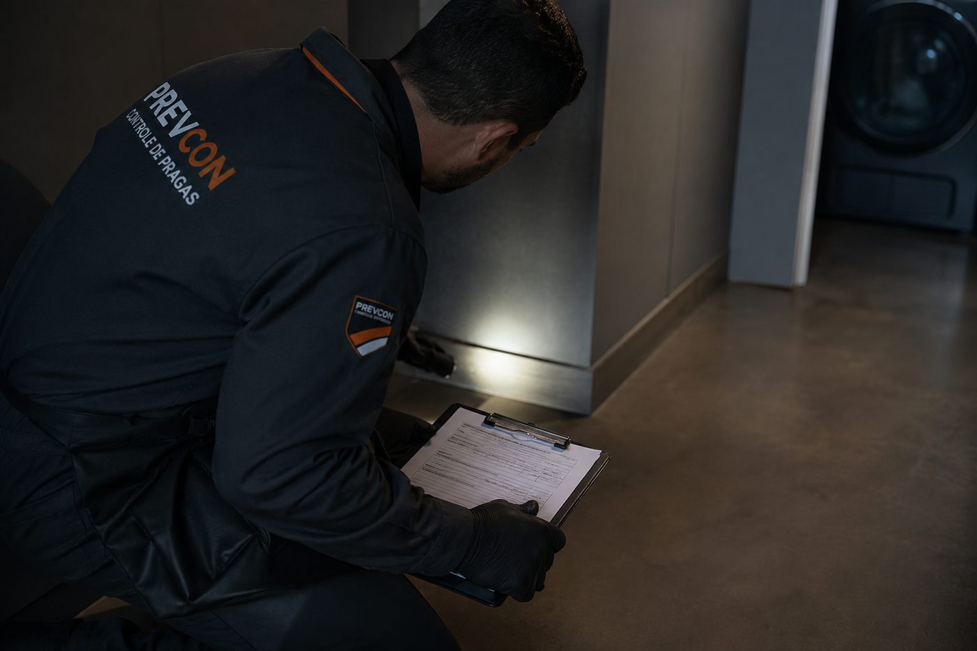Profissional de controle de pragas inspecionando o rodapé de uma área de serviço